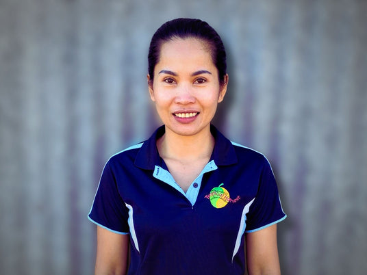 Ati, Processing Room Leader at Tropical Harvest, wearing uniform and smiling in front of tin shed wall