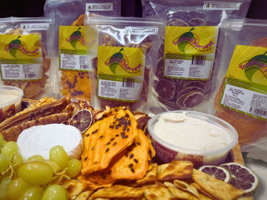 Tropical Harvest premium Australian dried fruit packs with pineapple, lime, and mango displayed alongside cheese, crackers, and grapes β a healthy, natural snack option
