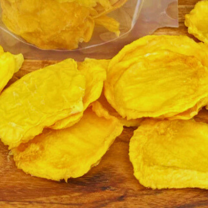 Green Dried Mango Minis shown on a wooden surface in front of clear packaging, highlighting their pale yellow colour and chewy texture.