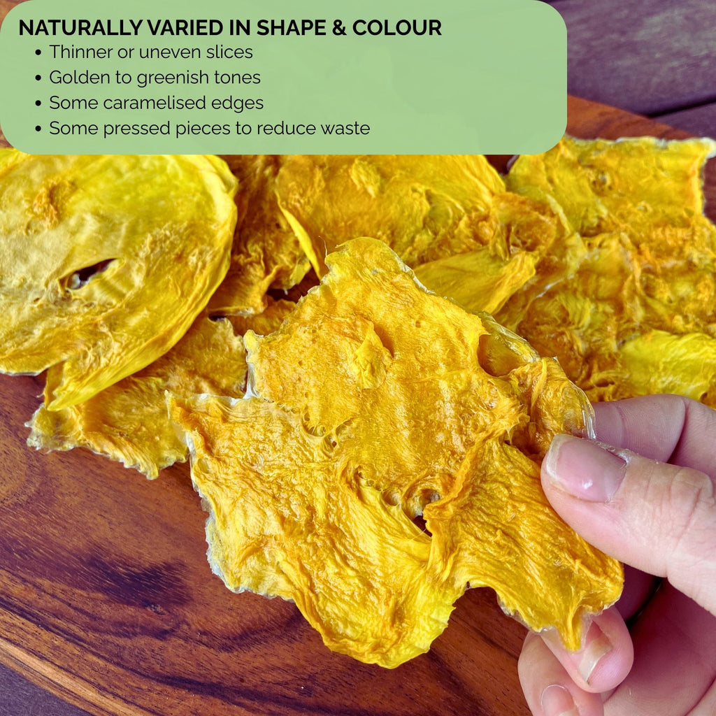 Close-up of Standard Australian Dried Mango slices on a wooden board, showing mixed shapes and tones. Text overlay reads “Naturally Varied in Shape & Colour – Thinner or uneven slices, golden to greenish tones, some caramelised edges, some pressed pieces to reduce waste