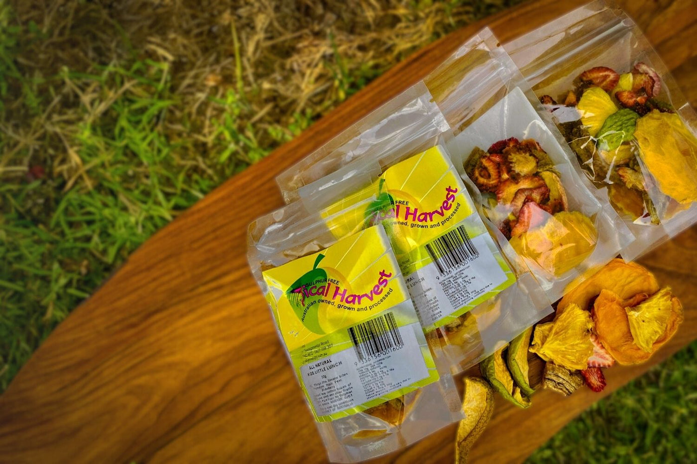 Tropical Harvest dried fruit packs with Australian mango, pineapple, banana, and strawberry slices displayed on a wooden table outdoors.